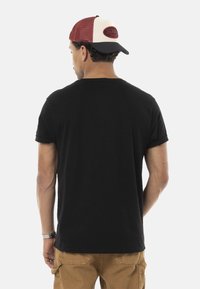 T-shirt noir à manches courtes avec un col rond et des poignets retroussés, porté avec un pantalon cargo beige. Une casquette de camionneur rouge et blanche est affichée à l'envers.