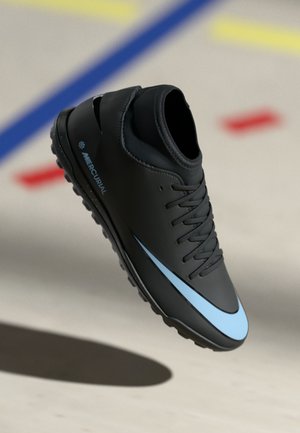 Black and blue athletic soccer shoe with laces and textured sole, hovering above a light-colored floor with red, blue, and yellow lines.
