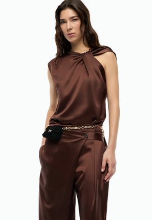 Donna che indossa un top marrone senza maniche in raso e pantaloni abbinati con cintura a catena dorata, tiene una piccola pochette nera, in piedi con le mani in tasca.