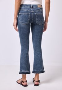 Jeans de mezclilla azules acampanados con una textura desteñida, diseño de cinco bolsillos y dobladillo sin terminar. La vista trasera muestra detalles de costura cerca del dobladillo.