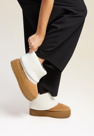 Chaussures à plateforme en suède beige avec une épaisse bordure en tricot côtelé blanc, portées avec un pantalon noir, mettant en valeur la texture et le design superposé.