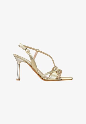 Sandalo dorato con tacco alto, sottili cinturini, decorazioni di strass, fibbia regolabile e punta quadrata aperta su sfondo bianco.