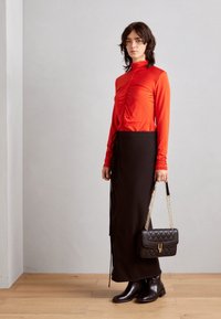 Czerwony top z długim rękawem i marszczeniami w połączeniu z czarną spódnicą do kostek z bocznymi wiązaniami. Model trzyma czarną pikowaną torebkę na łańcuszku w kolorze złotym.