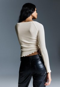 Kremowa, prążkowana bluzka z długim rękawem, z marszczeniem po bokach i falbaniastym dołem, zestawiona z czarnymi spodniami z ekoskóry z tylnymi kieszeniami.