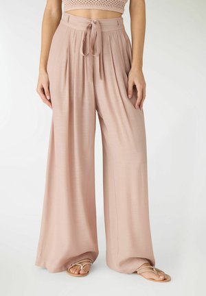 Donna che indossa pantaloni rosa cipria a vita alta con gambe larghe e cintura annodata, abbinati a sandali infradito beige e maglia corta a maglia.