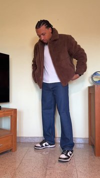 Casaco de fleece castanho com um colarinho alto, t-shirt branca por baixo, jeans azuis e ténis pretos com detalhes brancos, de pé no interior.