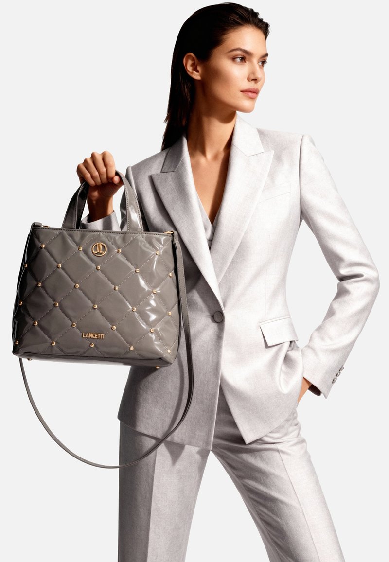 Borsa a mano grigia in vernice con design trapuntato, dettagli in metallo dorato, manici superiori e tracolla rimovibile. Logo visibile sulla parte anteriore.