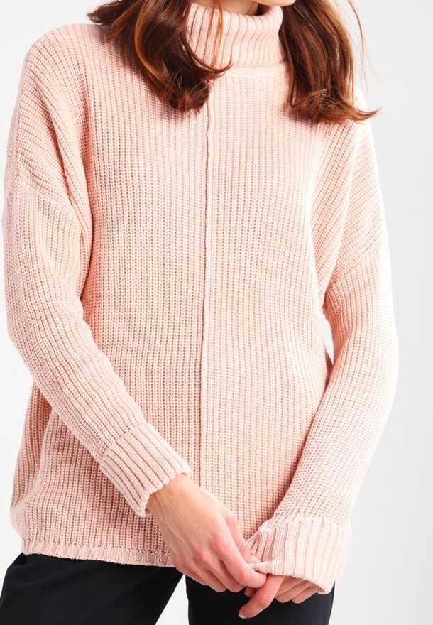Femme portant un pull col roulé en maille côtelée rose clair, tenant le poignet de la manche gauche avec les deux mains.