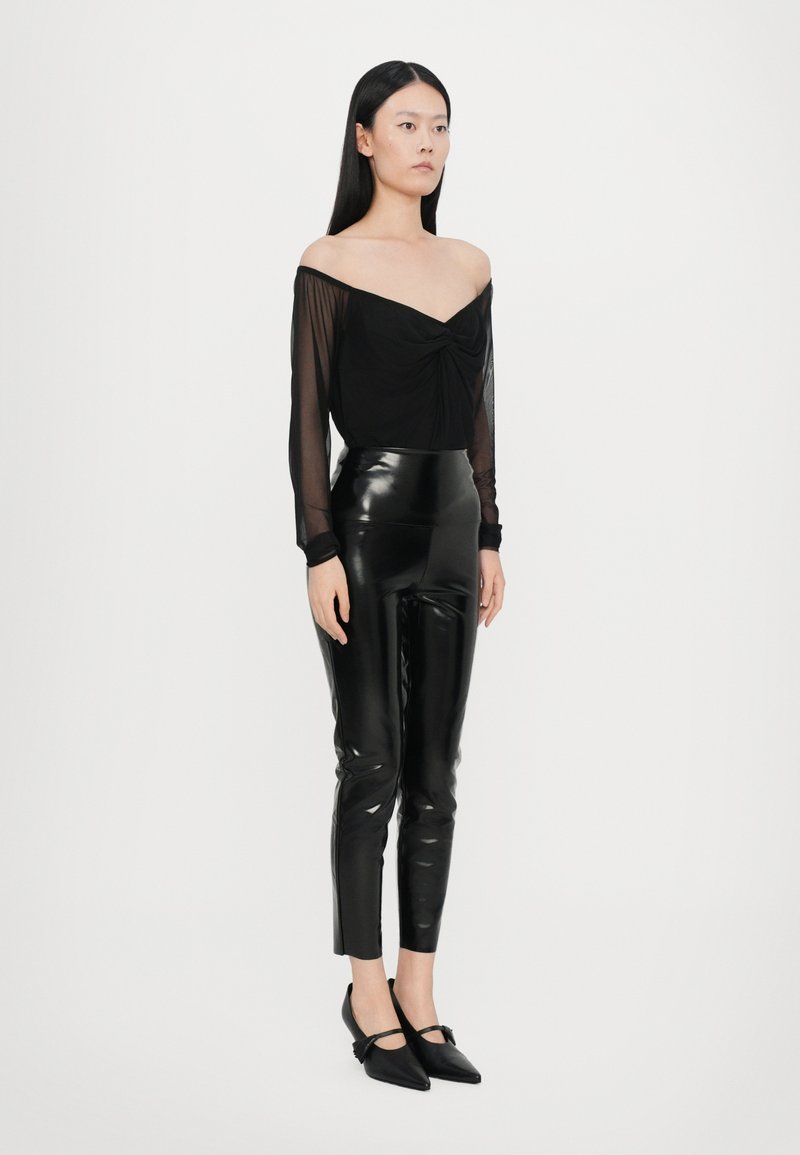 Top nero scollato con dettagli annodati, abbinato a pantaloni in lattice neri a vita alta e scarpe ballerine a punta con un accento a fiocco.