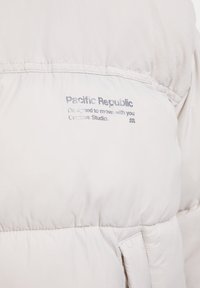 Jasnoszara kurtka puchowa z miękką teksturą, przeszyciami oraz metką z napisem "Pacific Republic" z tyłu.