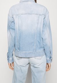 Giacca di jeans azzurro chiaro con effetto sbiadito. Presenta un colletto classico, due tasche posteriori e polsini con bottoni. Sono visibili dettagli strappati.