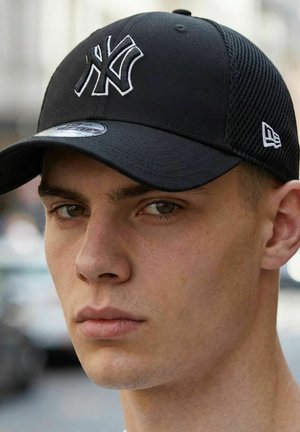 Junger Mann mit schwarzer New York Yankees Mütze mit weißem Logo, schaut frontal mit neutralem Gesichtsausdruck, unscharfer urbaner Außenhintergrund.