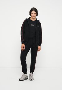 Sudadera con cremallera negra y franjas laterales rojas, camiseta negra y pantalones de chándal a juego; zapatillas gris con suelas negras; textura de tela suave.