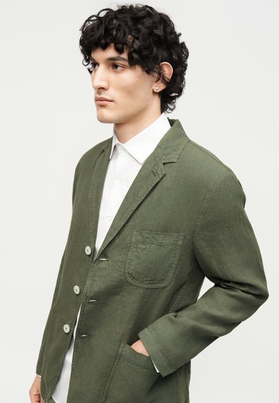 Νέος άνδρας με σγουρά σκούρα μαλλιά, που φοράει ένα χακί πράσινο σακάκι με κουμπιά πάνω από ένα λευκό πουκάμισο με γιακά, χέρι στην τσέπη, κοιτάζει αριστερά.