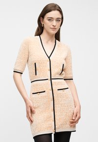 Νεαρή γυναίκα που φοράει πλεκτό φόρεμα σε ροδακινί απόχρωση με μαύρες λεπτομέρειες, κουμπιά στο μπροστινό μέρος και τέσσερις τσέπες, στέκεται μπροστά σε απλό φωτεινό φόντο.