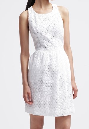 Robe blanche sans manches en tissu ajouré avec une taille cintrée et une jupe longueur genoux, portée par une personne aux longs cheveux foncés.