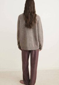 Grau gestrickter Pullover mit weiten Ärmeln, kombiniert mit lockeren lila Hosen. Der Hintergrund ist neutral mit einer sanften Teppichtextur.