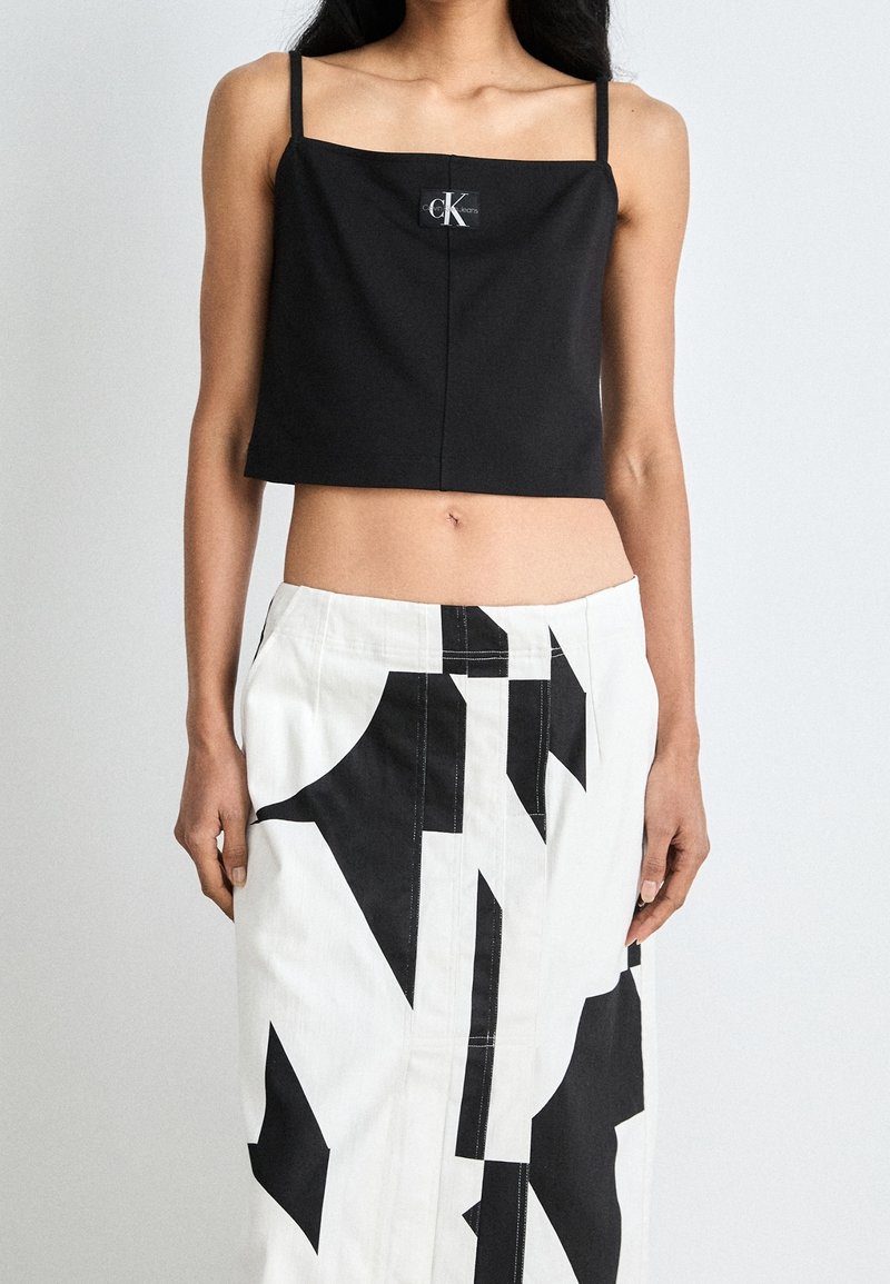 Femme portant un caraco noir raccourci avec le logo Calvin Klein et une jupe blanche taille haute avec des motifs abstraits noirs audacieux.