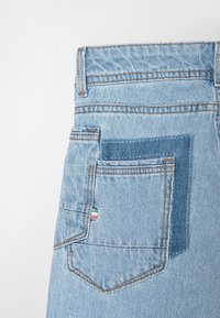 Jean en denim bleu clair avec une poche à patch accentuée d'un bleu plus foncé et une petite étiquette colorée en rouge, blanc et vert sur le côté.