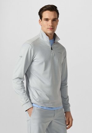 Hombre con camiseta de media cremallera gris claro y pantalones con las manos en los bolsillos, de pie frente a un fondo claro y liso.