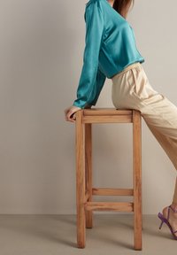 Tealfärgad satin långärmad topp som matchas med krämfärgade byxor, sitter på en träbarstol. Har lavendelfärgade remsandaler och släta texturer.