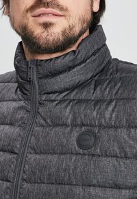 Chaqueta de plumas gris carbón con cuello alto, cierre de cremallera frontal y un parche de logo de goma en el lado izquierdo. Tejido texturizado y ligero.