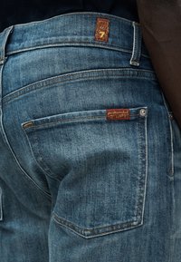 Μπλε τζιν παντελόνι με ταμπέλα από σκούρο καφέ δέρμα στην πίσω τσέπη. Διαθέτει ραφές λεπτομέρειας και σχεδιασμό με ίσια πόδια.