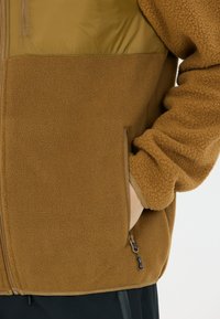 Ruskea fleece-takki, jossa on sileä oliivinvihreä olkapaneeli, sivu- ja vetoketjupussit sekä joustavat hihansuut tiiviiseen istuvuuteen.