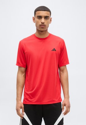 Hombre con camiseta deportiva roja de manga corta Adidas y pantalones cortos negros con patrones de chevrones blancos, de pie sobre un fondo liso.