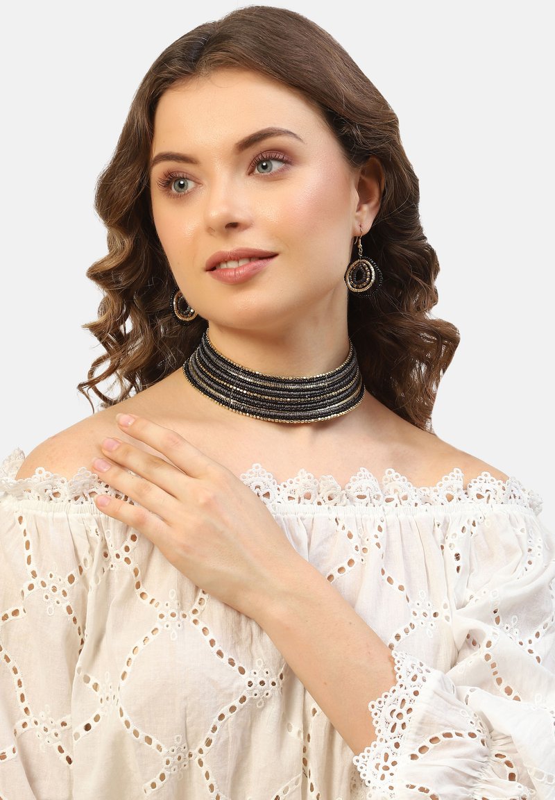Svart pärlhalsband med guldaccenter, parat med matchande hoops örhängen. Modellen bär en off-shoulder vit blus med spetsdetaljer.