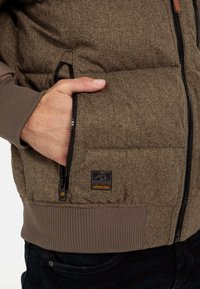 Brun oppustelig jakke med herringbone mønster, lynlåslommer og ribbede manchetter. Logoen viser hænder og teksten "MADE FOR DAILY ADVENTURES."