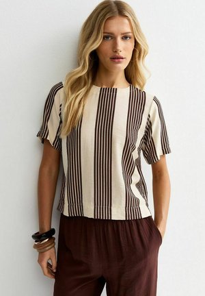 Donna bionda che indossa una maglietta a maniche corte beige e marrone con righe verticali, pantaloni marrone scuro e braccialetti impilati sul polso sinistro.