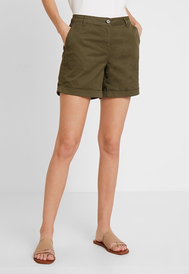 Shorts en coton vert olive avec une fermeture à bouton, deux poches latérales, ourlets retroussés, coupe droite, associés à des sandales plates couleur beige.