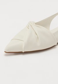 White flat shoe made of smooth material with a pointed toe, featuring a twisted design detail and a cut-out side for easy wear.