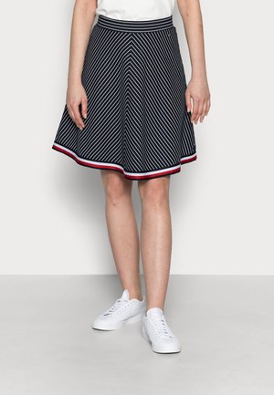 Vrouw draagt een marineblauwe wijde rok met witte diagonale strepen en rode, witte en blauwe rand, gecombineerd met witte sneakers.