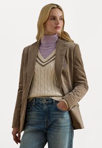 Blazer di velluto a coste marrone con rever a notched, abbinato a un maglione a treccia crema e a un dolcevita viola chiaro, indossato con jeans in denim blu.