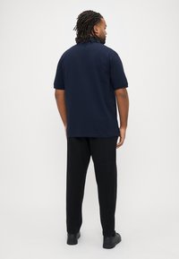 Marineblå polo med korte ærmer, sideslidser og krave, kombineret med sorte bukser og sorte sko. Set bagfra mod en ensfarvet baggrund.