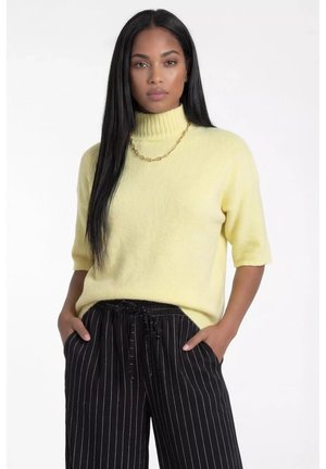 Kobieta o długich czarnych włosach nosi żółty sweter z krótkim rękawem i golfem, złoty łańcuszek oraz czarne spodnie w cienkie prążki, z rękami w kieszeniach.