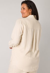 Long-sleeve, beige shirt with a relaxed fit, back pleat, and curved hem. Fabric has a soft texture and a natural, lightweight appearance.