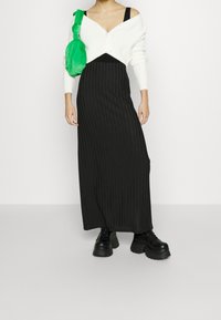 Frau trägt einen schwarzen gerippten Maxirock, ein schwarzes Oberteil, eine weiße kurze Strickjacke, schwarze Plateaustiefel und eine leuchtend grüne Umhängetasche.