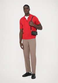 Polo rojo de manga corta con cremallera, pantalones beige a medida y zapatillas negras. Además, se sostiene una pequeña bolsa negra con correa en una mano.