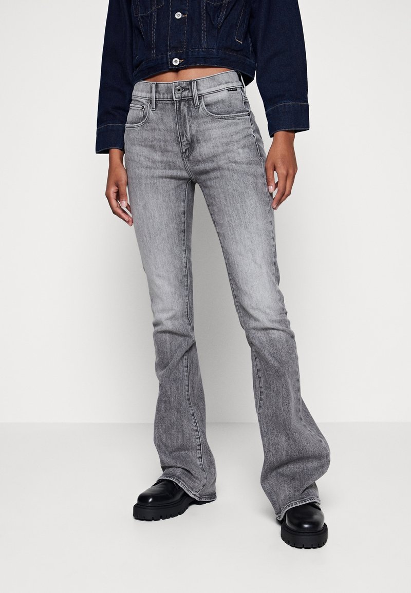 Personne portant un jean bootcut gris délavé, des chaussures noires épaisses et une veste courte en jean foncé, debout devant un fond clair uni.