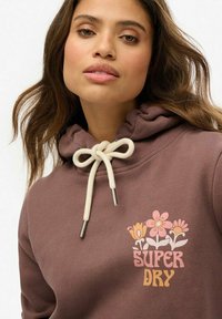 Hoodie marron avec un cordon de serrage, ornée d'un imprimé floral et du texte "SUPER DRY" dans des couleurs variées. Texture douce et design décontracté.