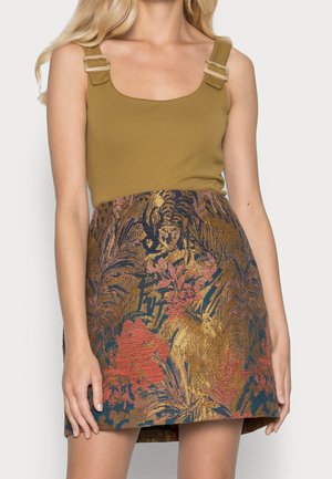 Kobieta w oliwkowozielonym topie na ramiączkach z klamrami oraz w spódnicy o wysokim stanie z brokatowym wzorem w czerwone i złote kwiaty.