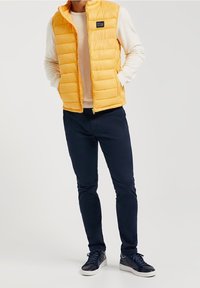 Gul pufferväst med dragkedja fram, randig textur, över en ljus topp, kombinerad med mörkblå byxor och marinblå sneakers. Enkel etikett på västen.