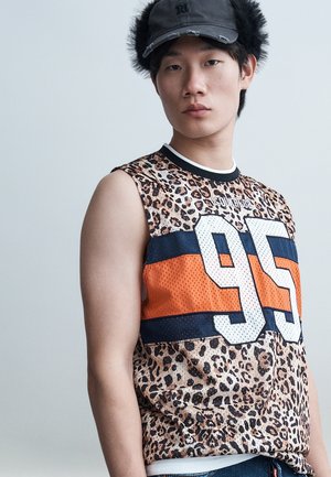 Jeune homme portant un débardeur à imprimé léopard avec un grand "95" ainsi que des rayures orange et marine, et une casquette noire avec des protège-oreilles en peluche.