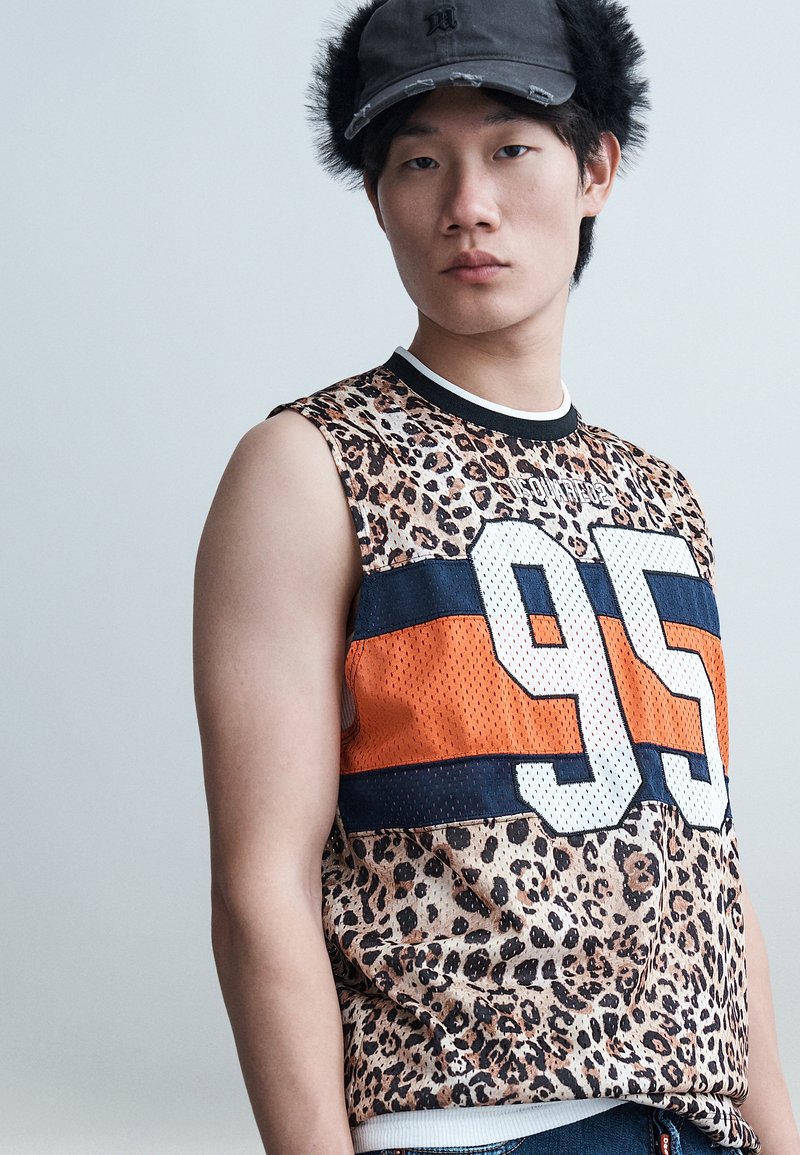 Jeune homme portant un débardeur à imprimé léopard avec un grand "95" ainsi que des rayures orange et marine, et une casquette noire avec des protège-oreilles en peluche.