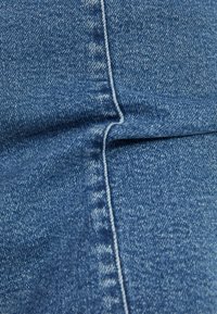 Blå denimtyg med en texturerad yta, synlig sömnad och en naturlig veck. Färgen är konsekvent, utan synliga mönster eller slitningar.