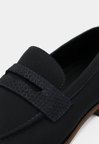 Μαύρο λουστρίνι με υφασμάτινη υφή και αντίθετη ραφή, με λείο πάνω μέρος. Διαθέτει λεπτό προφίλ και ξύλινη σόλα.