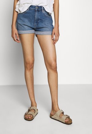 Person iført rullede denimshorts, beige slip-on sandaler med spænder og en hvid top med blondedetaljer, stående mod en ensfarvet baggrund.
